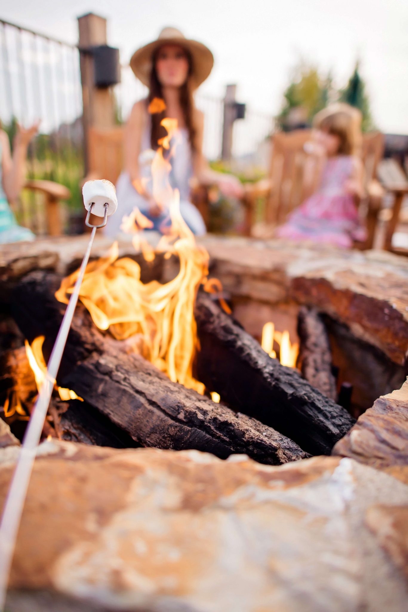 A marshmallow roasting over a stone fire pit, family gathered on a porch in the background